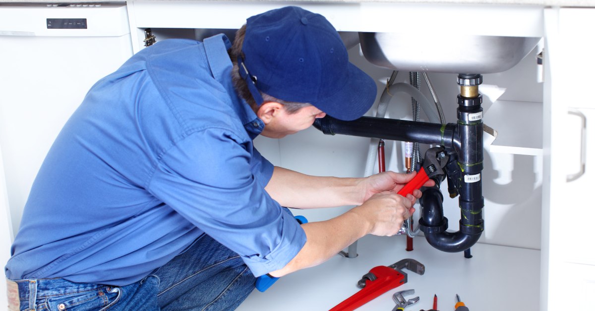 A plumber repairs copper pipes with a wrench while kneeling on the floor near exposed plumbing connections.