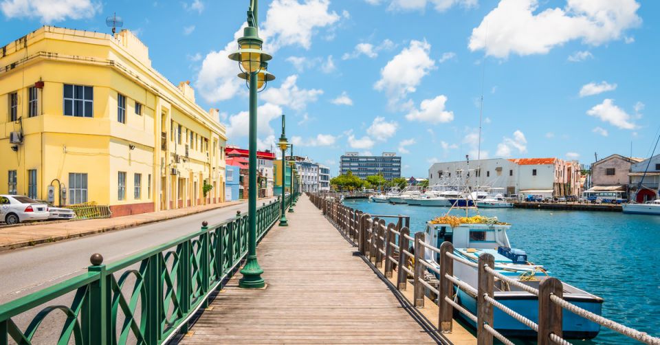 An empty marina promenade in Barbados stretches under a blue sky, surrounded by colorful buildings and small boats.
